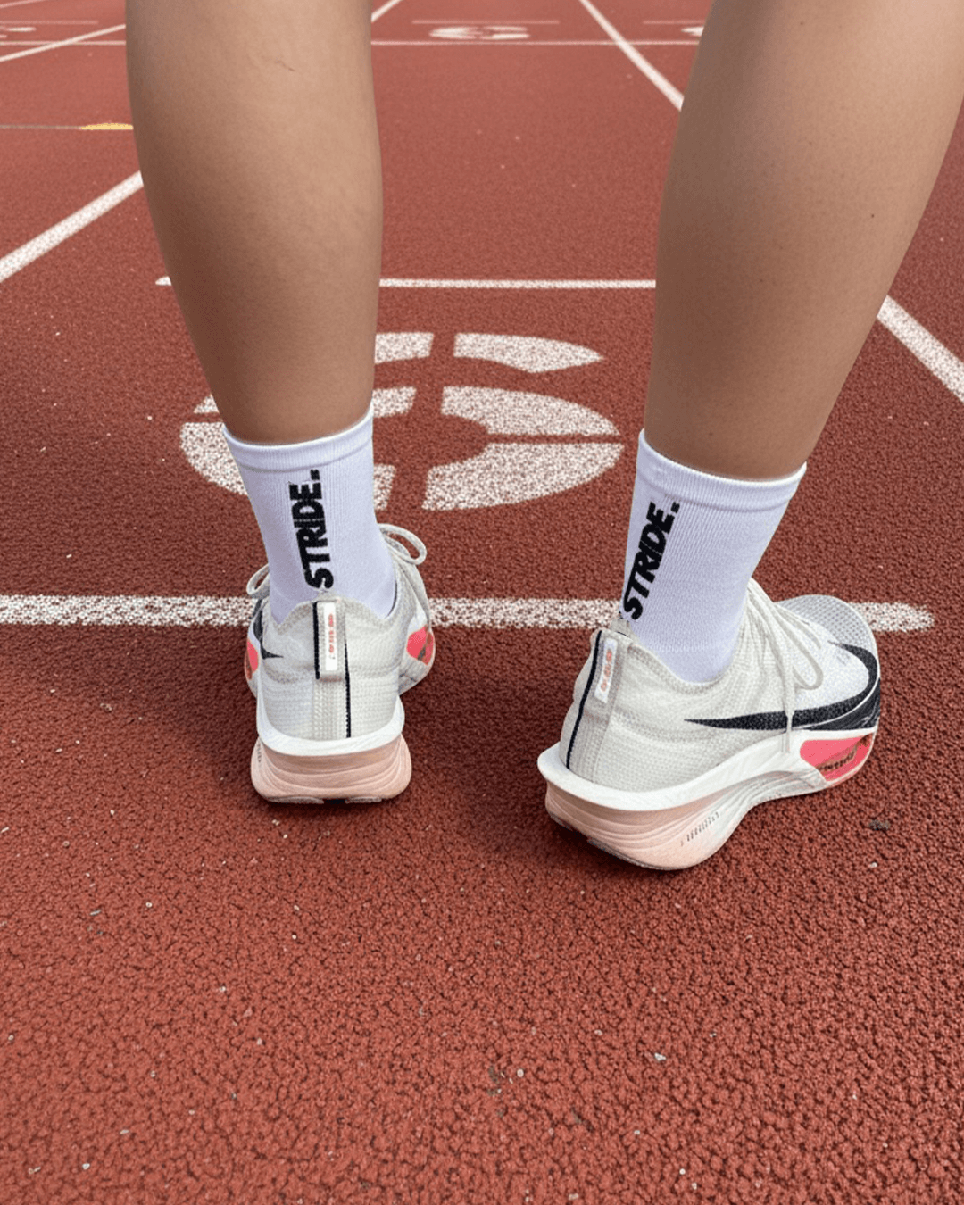 A person's legs and feet wearing white socks and running shoes, standing on a track.