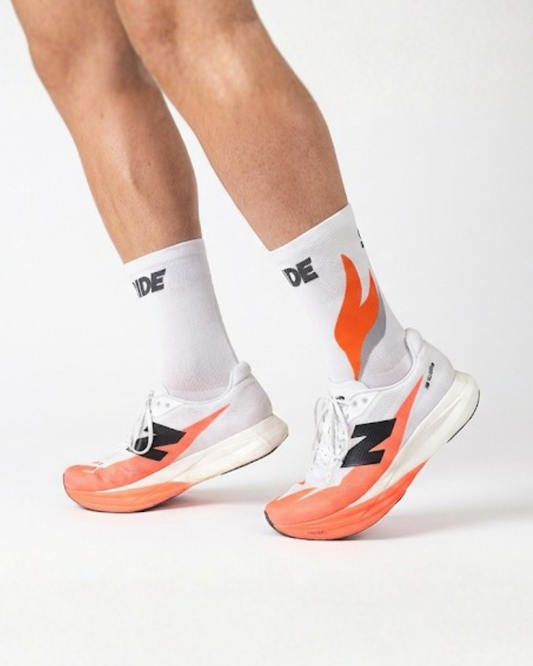 White sneakers with orange soles and black accents worn with white socks featuring a logo.